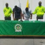 En la imagen se observa a un hombre de camiseta azul oscura y a otro hombre de camiseta blanca, custodiados por dos agentes de la Policía Nacional, delante de ellos se encuentra una mesa donde se evidencian armas de fuego y municiones.
