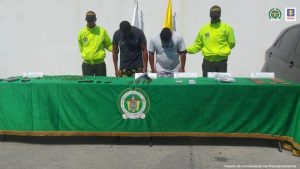 En la imagen se observa a un hombre de camiseta azul oscura y a otro hombre de camiseta blanca, custodiados por dos agentes de la Policía Nacional, delante de ellos se encuentra una mesa donde se evidencian armas de fuego y municiones.