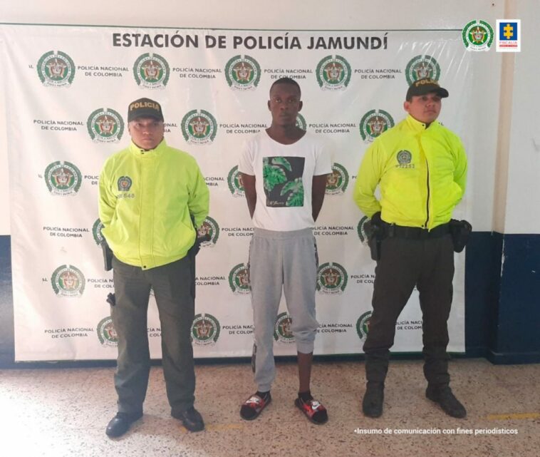 En la foto aparecen, de pie y con los brazos atrás, Jhon Bayron Cuero Sepúlveda. Viste camiseta blanca de manga corta con estampado; sudadera gris y pantuflas. A lado y lado de él hay uniformados de la Policía Nacional. Detrás de ellos hay un pendón institucional de la Policía.