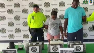 En la fotografía se observa a una mujer morena con camiseta gris, licra rosada esposada junto a un hombre de camiseta azul clara con bermuda azul oscura, custodiado por un agente de la Policía Nacional. Igualmente se muestra en una mesa la droga incautada.