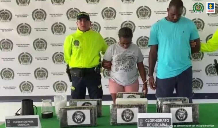 En la fotografía se observa a una mujer morena con camiseta gris, licra rosada esposada junto a un hombre de camiseta azul clara con bermuda azul oscura, custodiado por un agente de la Policía Nacional. Igualmente se muestra en una mesa la droga incautada.