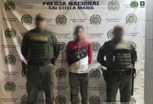 En la imagen se observa a  un hombre de camiseta roja con rallas azul oscura y blanca con un jean negro custodiado por dos agentes de la Policía Nacional delante de un pendón de esa institución.