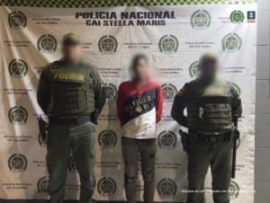 En la imagen se observa a  un hombre de camiseta roja con rallas azul oscura y blanca con un jean negro custodiado por dos agentes de la Policía Nacional delante de un pendón de esa institución.