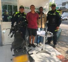 En la fotografía se identifica a dos hombres, uno con playera roja, short negro y tenis negros y el otro con playera amarilla, jeans azules y tenis blancos, custodiados por dos agentes de la Policía Nacional.  También hay una mesa frente a ellos con el arma que le fue incautada durante las detenciones.