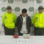 En la foto se le capta con dos policías nacionales.  Al fondo, hay una mesa con un frasco de plástico y una bolsa de morfina.