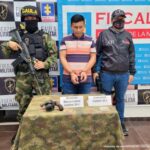 capturado esposado de frente custodiado por investigador del CTI de la Fiscalía y militar del Ejército Nacional. Delante de ellos una mesa con arma y munición incautaos al detenido, detrás banner de fiscalía y de Gaula Militar.