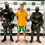 En la imagen se observa a un hombre de tez blanca vestido con camiseta amarilla, bermuda verde y sandalias negras, custodiado por dos agentes de la Policía Nacional delante de un pendón de esa institución.