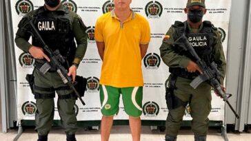 En la imagen se observa a un hombre de tez blanca vestido con camiseta amarilla, bermuda verde y sandalias negras, custodiado por dos agentes de la Policía Nacional delante de un pendón de esa institución.