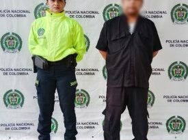 En la imagen se observa a un hombre de tez blanca vestido con pantalón, camisa y zapatos negros, custodiado por una uniformada de la Policía Nacional, delante de un pendón de esa institución.
