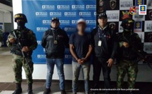 Capturado con gorra de frente esposado, custodiado por dos investigadores del CTI de la Fiscalía y dos militares del Ejército Nacional, detrás banner de Fiscalía y Ejército Nacional.