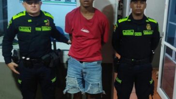 En la fotografía aparece un  hombre moreno de camiseta roja, con bermuda de jean, custodiado por dos agentes de la Policía Nacional en una estación de esa institución.