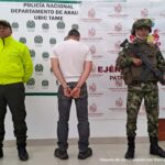 En la imagen se observa al capturado de espaldas junto a dos uniformados de la Policía y Ejército Nacional.  Detrás de ellos los banners que identifican a la Departamento de Policía de Tame y al Ejército Nacional.