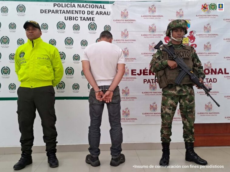 En la imagen se observa al capturado de espaldas junto a dos uniformados de la Policía y Ejército Nacional.  Detrás de ellos los banners que identifican a la Departamento de Policía de Tame y al Ejército Nacional.