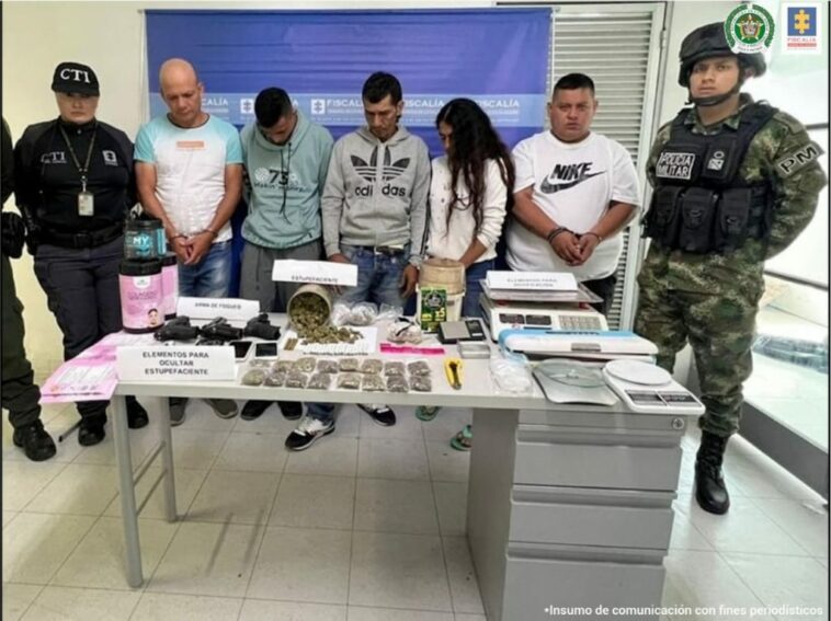 En la foto aparecen los cinco presuntos integrantes del grupo delincuencial Los de Nápoles, entre ellos una mujer. A lado y lado de ellos hay uniformados de la Policía Nacional. Frente a ellos hay un escritorio gris y sobre él hay recipientes con etiquetas alusivas a productos de belleza; bolsas plásticas y tarros que contienen material vegetal, armas de fuego y grameras. Detrás de ellos hay un pendón institucional de la Fiscalía.