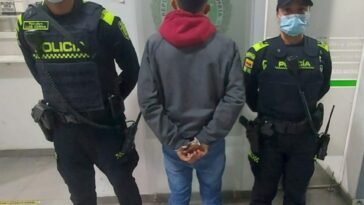 Capturado de espaldas esposado, custodiado por dos uniformados de la Policía. Detrás aviso de Policía Nacional en terminal de transporte.