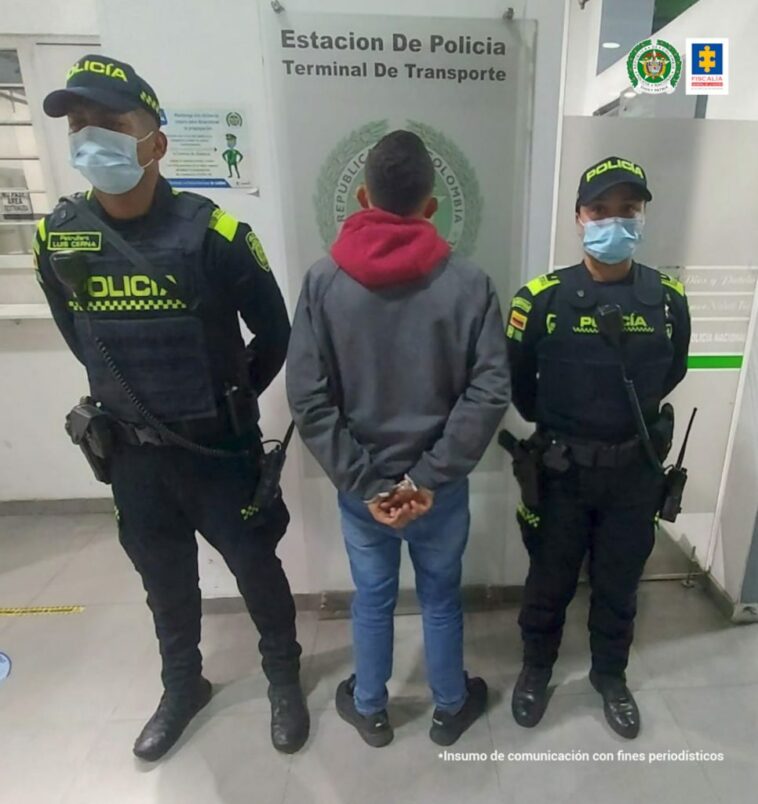 Capturado de espaldas esposado, custodiado por dos uniformados de la Policía. Detrás aviso de Policía Nacional en terminal de transporte.