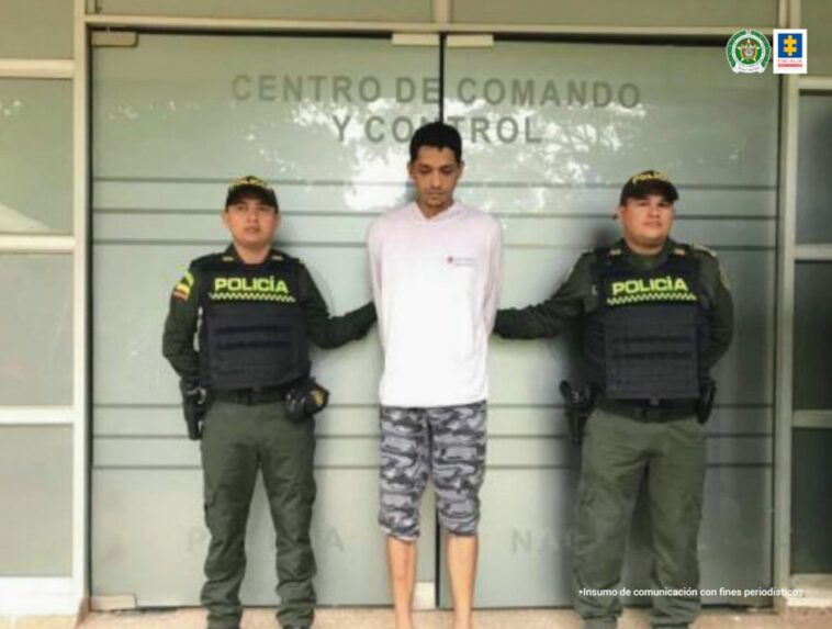 capturado de frente en pantaloneta y esposado manos atrás, custodiado por dos uniformados de la Policía Nacional. Detrás banner de la Policía Nacional.