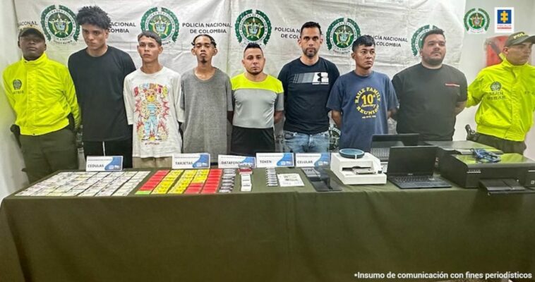 Dos uniformados de la Policía Nacional custodian a los procesados quienes fueron capturados en los barrios Villa Campestre y Las Delicias de la ciudad de Barranquilla. En la mesa están los elementos incautados como computadores, celulares, datáfonos inalámbricos, tokens, tarjetas débito, tarjetas de créditos, cédulas de ciudadanía colombiana, licencias de conducción de los Estados Unidos, documento de identificación de España, impresoras