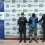 capturado de frente cabeza agachada esposado manos atrás, custodiado por dos uniformados de la policía Nacional. Detrás banner de la Policía Nacional