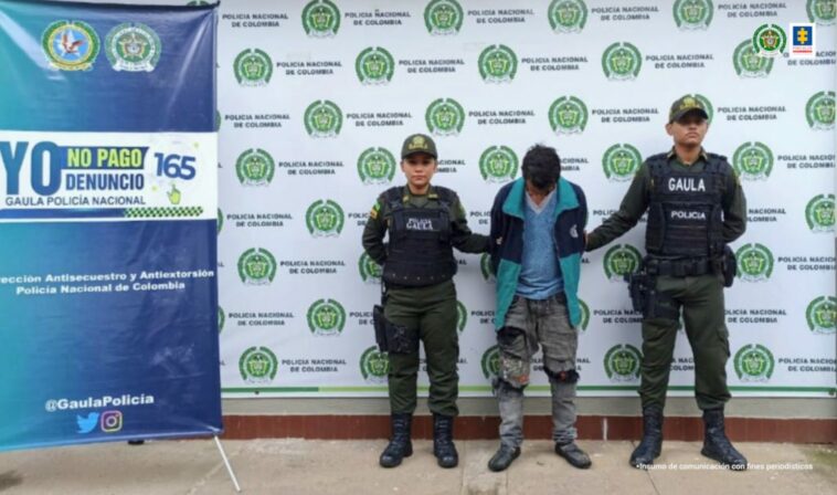 capturado de frente cabeza agachada esposado manos atrás, custodiado por dos uniformados de la policía Nacional. Detrás banner de la Policía Nacional