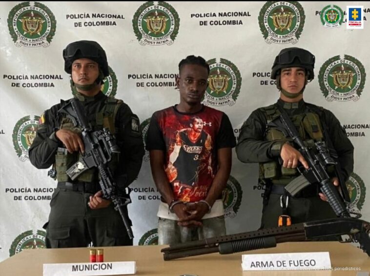 En la fotografía aparece el capturado junto a dos agentes de la Policía Nacional. En la parte posterior está una mesa con un arma de fuego y munición .