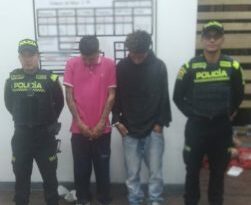 los dos hombres estan acompañados de dos uniformados de la Policía Nacional delante de un pendon de la metropolitana de Bogotá.