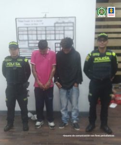 los dos hombres estan acompañados de dos uniformados de la Policía Nacional delante de un pendon de la metropolitana de Bogotá.