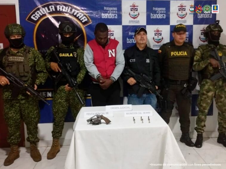 En la fotografía aparece el capturado junto a un servidor del CTI, un agente de la Policía, uniformados del Gaula Militar y la Armada Nacional. En la parte superior está una mesa con un arma de fuego y munición 