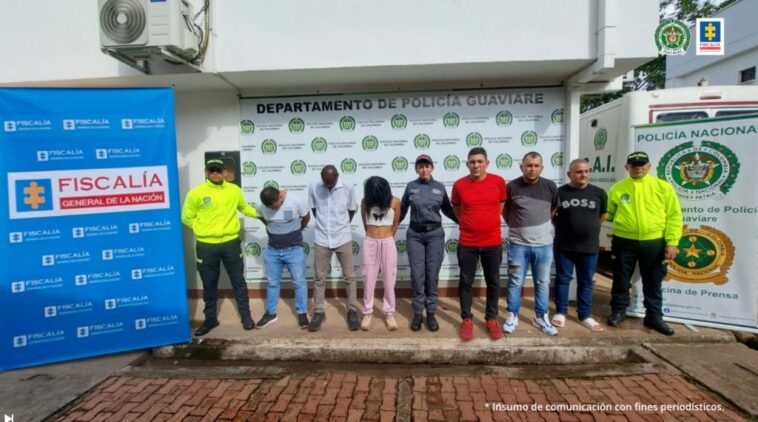 están los capturados de f4ente con manos esposadas atrás, custodiados por dos uniformados de a Policía Nacional detrás banner de la Policía Nacional.