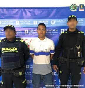 En la fotografía aparece un hombre moreno de pie, vestido con camiseta blanda de rallas azules y gris, jean azul, custodiado por dos uniformados de la Policía Nacional. Detrás de ellos hay un pendón azul que dice Fiscalía General de la Nación.