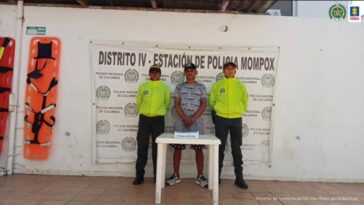 En la imagen aparece un hombre moreno con los brazos adelante vestido con gorra, camiseta y bermuda de colores blanco con negro y unos tenis de color negros. Se encuentra custodiado por dos agentes de la Policía Nacional y detrás de ellos está un pendón que dice Distrito IV Estación de Policía Mompox.