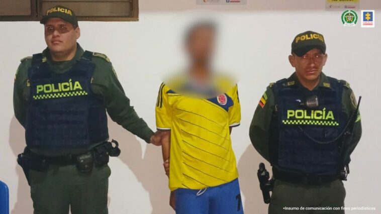 El capturado viste una camiseta de la selección Colombia, está esposado con sus manos a la espalda y está custodiado por dos uniformados de la policía nacional.