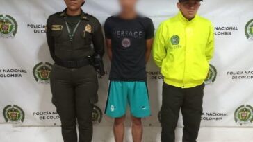 El capturado está en medio de dos uniformados de la policía nacional, tiene una pantaloneta verde y una camiseta negra y sus manos esposadas a a la espalda.