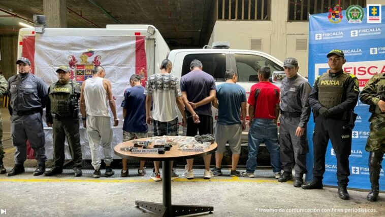 Se observan seis personas capturas, en fila y de espaldas.  En casa costado uniformados del CTI y de Ejército Nacional. Al fondo un pensón de Fiscalía General de la Nación y uno de Ejército Nacional, así como una unidad móvil.  Al frente una mesa con material incautado en las diligencias de allanamiento