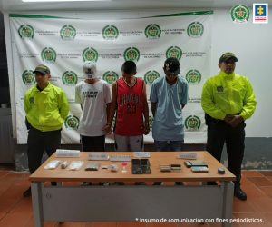 capturados de frente custodiados por policías. Delante mesa con material incautado, Detrás banner de la Policía Nacional.