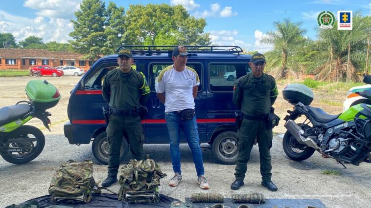 está el capturado de frente manos atrás esposado custodiado por dos policías, delante n el piso material incautado. Detrás camioneta también incautada.