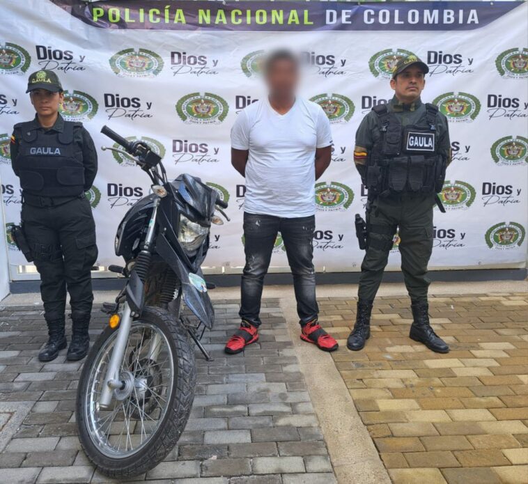 capturado de frente esposado custodiado por dos policías del grupo Gaula, al lado motocicleta incautada. Detrás banner de la Policía Nacional.