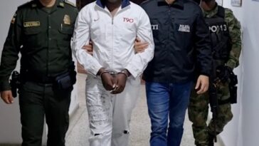 En la fotografía se aprecia, en acción de caminata y esposado a Jhoason Estiven Camacho Mosquera. Viste camisa de manga larga blanca; pantalón y tenis blancos. Al lado izquierdo hay un servidor del CTI de la Fiscalía y un uniformado del Ejército. Al lado derecho hay un uniformado de la Policía Nacional.