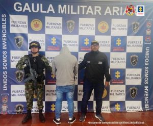 Capturado de espaldas con saco de capucha que le cubre la cabeza, de espaldas manos adelante, custodiado por un investigador del CTI de la Fiscalía y un militar. Detrás banner de Gaula Militar y Fiscalía.