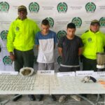 En la fotografía se aprecia a dos de los tres presuntos implicados en el hurto a la fundación de seguidores de un equipo de fútbol. Uno viste buzo de sudadera con franjas azul, blanca y negra y la marca ‘Puma’ en el torso; pantaloneta camuflada y pantuflas. La otra persona viste camisa de la Selección Colombia azul; jean con desgaste y zapatillas. A lado y lado de ellos hay uniformados de la Policía Nacional y detrás de ellos hay un pendón institucional de la Policía.