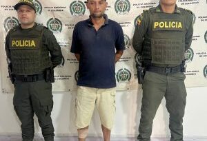 En la imagen se ve una persona detenida bajo custodia de dos integrantes de la Policía Nacional. Detrás suyo un backing institucional.