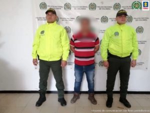 en la imagen se ve un hombre detenido bajo custodia de dos uniformados de la Policía Nacional. Detrás suyo un backing institucional.