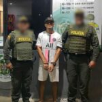 En la fotografía se encuentra un hombre de pie, vestido de camiseta y bermuda blanca, con sandalias de rallas negras, blancas y rojas. Sus manos están esposadas y aparece acompañado de dos uniformados de la Policía Nacional.