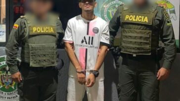 En la fotografía se encuentra un hombre de pie, vestido de camiseta y bermuda blanca, con sandalias de rallas negras, blancas y rojas. Sus manos están esposadas y aparece acompañado de dos uniformados de la Policía Nacional.