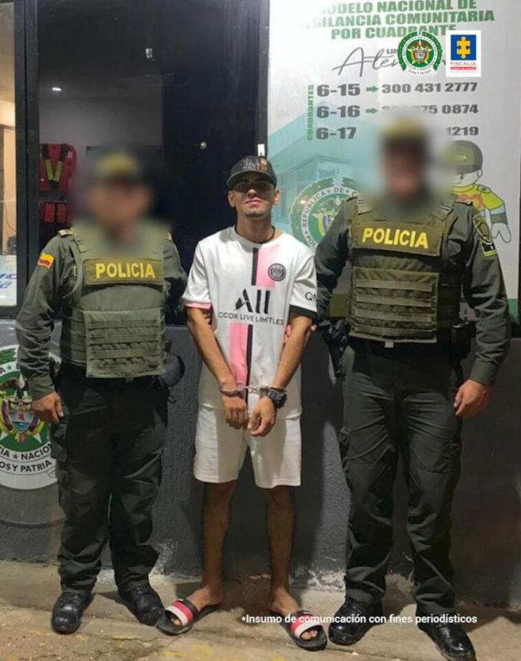 En la fotografía se encuentra un hombre de pie, vestido de camiseta y bermuda blanca, con sandalias de rallas negras, blancas y rojas. Sus manos están esposadas y aparece acompañado de dos uniformados de la Policía Nacional.