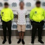 En la fotografía hay un hombre moreno de pie, con los brazos atrás, vestido con camiseta blanca, bermuda beis, zapatos tenis negros, custodiado por dos agentes de la Policía Nacional. Detrás de ellos hay un pendón de esa institución.