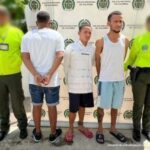 En la fotografía aparecen tres hombres vestidos de camisetas blancas con bermudas, todos se encuentran de pie, custodiados por dos agentes de la Policía Nacional. Detrás de ellos hay un pendón de esa institución.