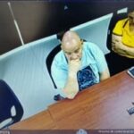 En la fotografía aparece un hombre sentado con los brazos sobre un escritorio de tez blanca, calvo y vestido de camiseta azul, con un micrófono en su boca.  A su lado se encuentra una mujer sentada de cabello negro, vestida con camiseta amarilla y pantalón negro