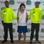 En la fotografía hay un hombre moreno, de pie, de cabellos crespos negro, vestido de camisilla blanca, con bermuda azul y sandalias blancas. Su brazos se encuentran tatuados y su pierna derecha también. Se encuentra custodiado por dos agentes de la Policía Nacional.