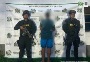 En la fotografía se visualiza  el capturado de espaldas junto a dos uniformados de la Policía Nacional. En la parte posterior se ubica el banner que identifica a la Policía Nacional.